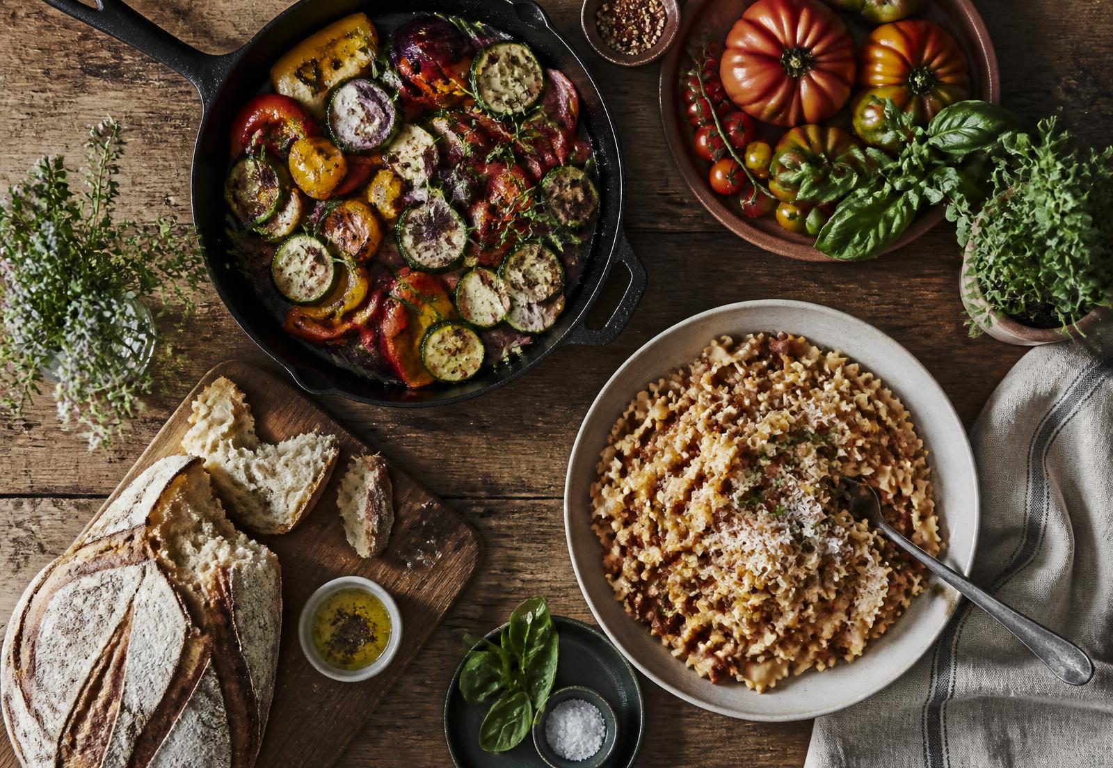 Overhead view of a wooden table set with roasted vegetables, sourdough and pasta